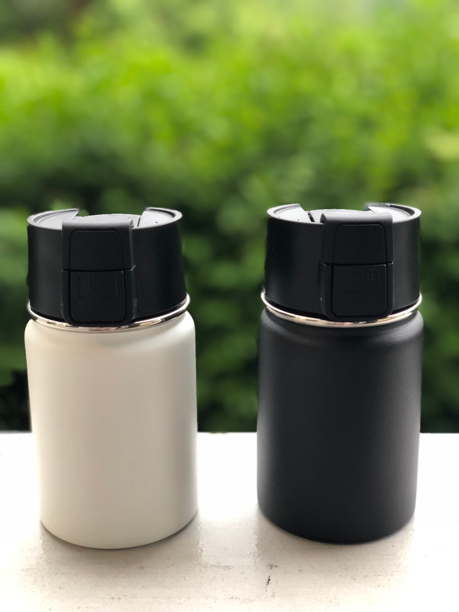 Weisser und schwarzer Kaffeebecher to go aus doppelwandigem Edelstahl mit Einhand Click-Verschluss 350 ML, dargestellt im Freien.