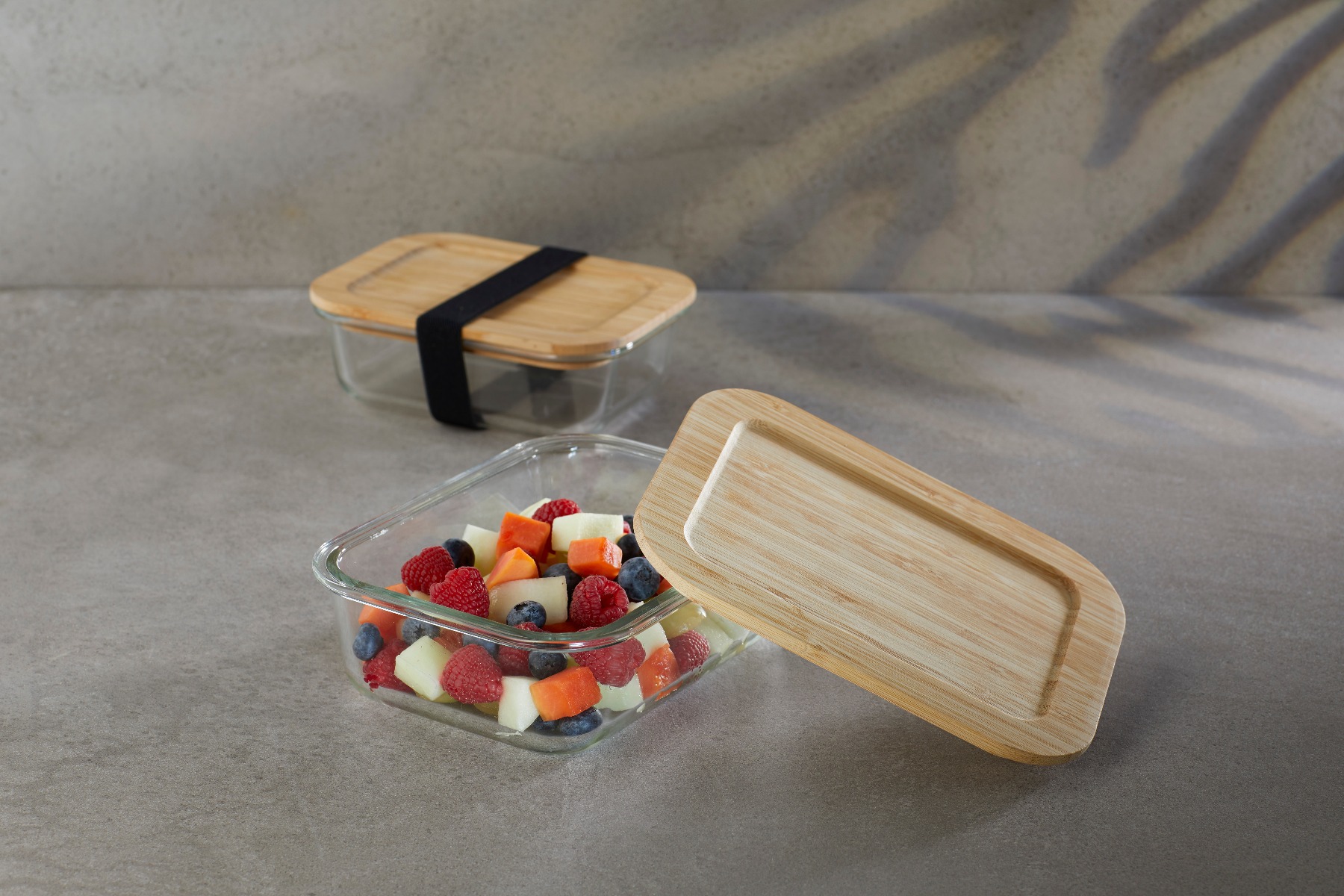 Umweltfreundliche Aufbewahrungsbox large aus hitzebeständigem Borosilikatglas mit schwarzem, elastischem Halteband, dargestellt mit und ohne Früchte.