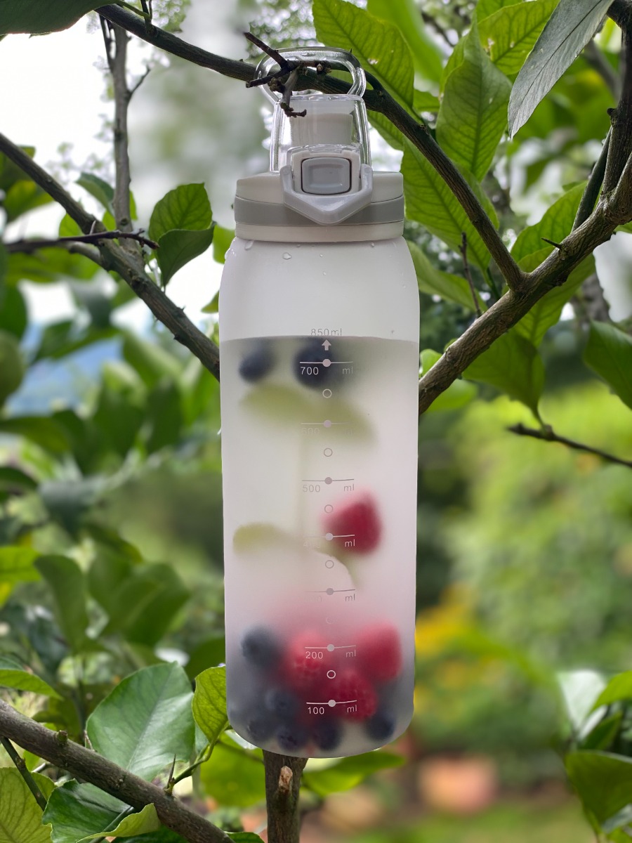 Trinkflasche aus gefrostetem Tritan, BPA frei, mit Klappdeckel und Sieb 900ML, dargestellt mit Flüssigkeit und Beeren, in einem Baum hängend.