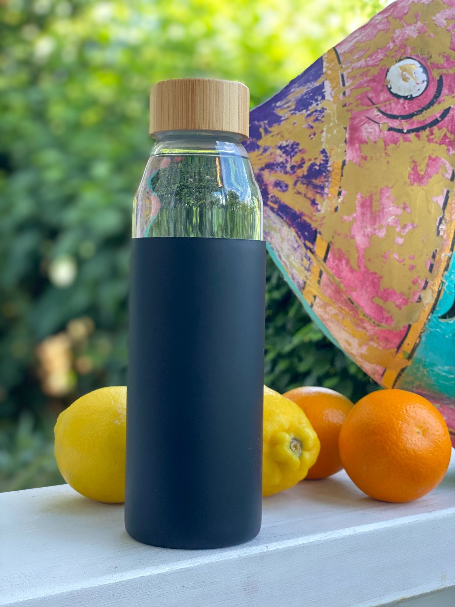 Umweltfreundliche Trinkflasche aus Glas mit schwarzer Silikonhülle und Bambusverschluss 500 ML, dargestellt im Freien mit Früchten im Hintergrund.