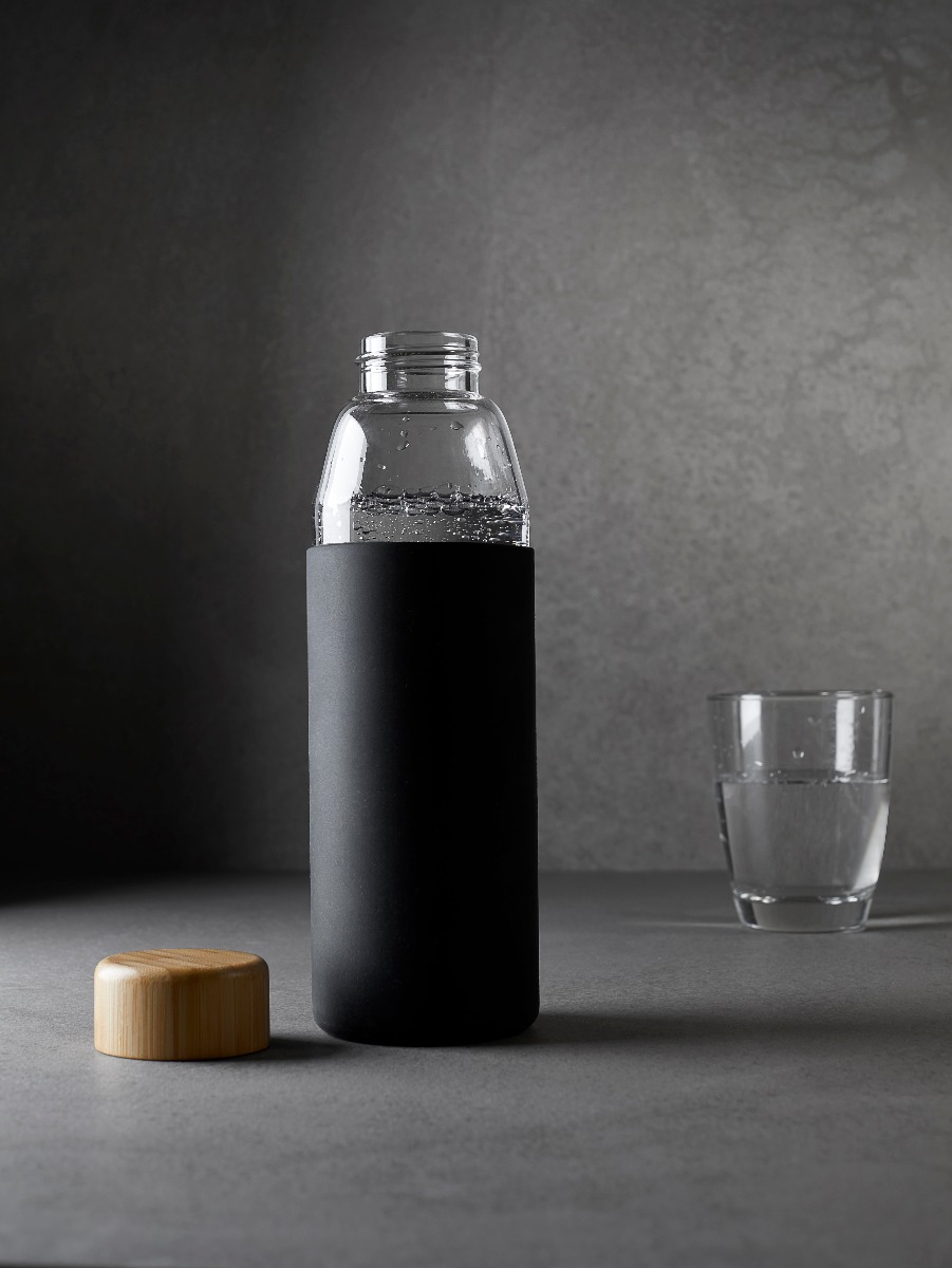 Umweltfreundliche Trinkflasche aus Glas mit schwarzer Silikonhülle und Bambusverschluss, dargestellt geöffnet und mit einem kleinen Glas an der Seite.