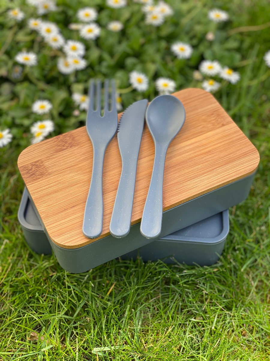 Umweltfreundliche Doppel-Lunchbox mit Bambusdeckel und Besteck, dargestellt auf einer Wiese, Besteck auf Deckel liegend.