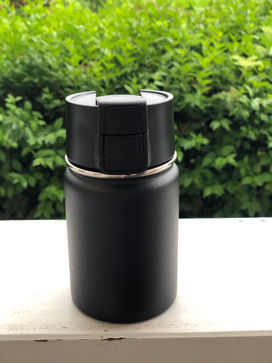 Schwarzer Kaffeebecher to go aus doppelwandigem Edelstahl mit Einhand Click-Verschluss 350 ML, dargestellt im Freien.
